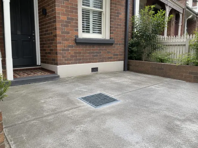Outdoor drain grate in a dry paved courtyard beside a brick house in Lilyfield